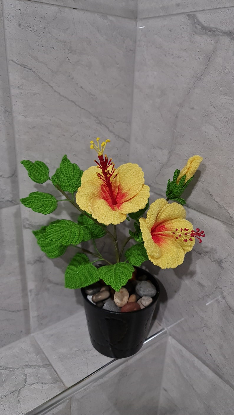 May include: A crocheted yellow hibiscus flower plant with red centres in a black pot with small stones. The plant is on a grey tiled surface.
