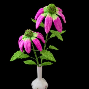 May include: Two handmade crochet flowers in a white vase against a black background. The flowers have bright pink petals with green tips and centers. Green leaves and stems support the flowers. The vase is white with a floral pattern.