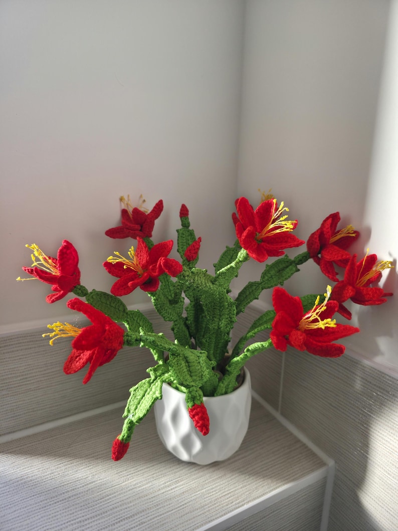 May include: A crocheted cactus plant with vibrant red flowers and yellow stamens, displayed in a white geometric pot. The green stems and leaves provide a striking contrast to the bright red blooms, creating a unique home decor item.