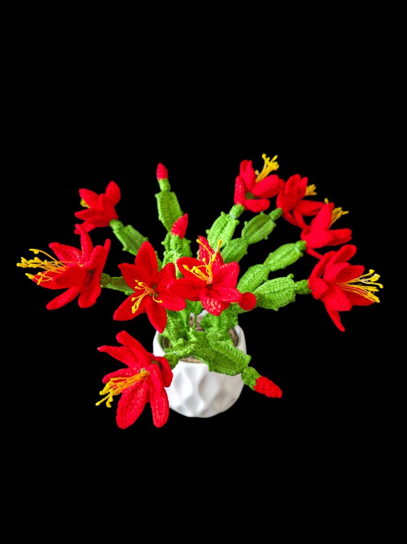 May include: A handmade crochet cactus plant with vibrant red flowers and yellow stamens, displayed in a white geometric pot. The green cactus stems and red buds add to the realistic look. The plant is set against a solid black background.