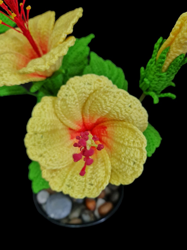 May include: A crocheted yellow hibiscus flower with a red centre, in a small pot with stones. The flower is in bloom and has a green stem and leaves.