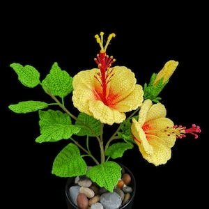 May include: A crocheted yellow hibiscus flower plant with red centres in a black pot with small grey and brown stones. The plant has green leaves.