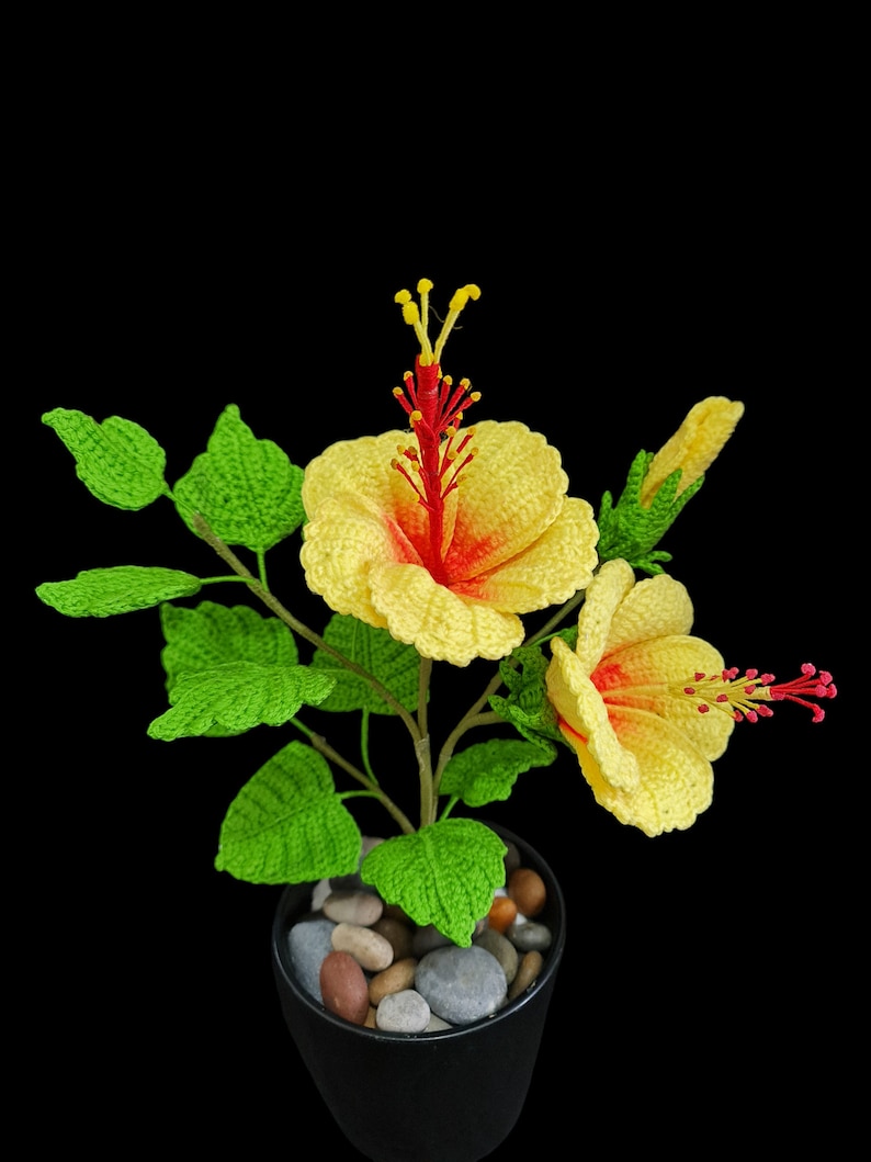 May include: A crocheted yellow hibiscus flower plant with red centres in a black pot with small grey and brown stones. The plant has green leaves.