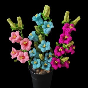 May include: A crocheted flower arrangement in a black pot. The flowers are pink, blue, and purple and have a bell shape. The pot is filled with small, smooth stones.