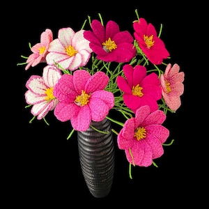 May include: A bouquet of pink and white crocheted cosmos flowers in a black vase. The flowers are arranged in a cluster, with the stems extending out from the vase.