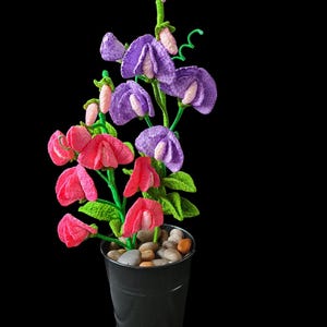 Könnte beinhalten: Eine Topfpflanze mit rosa und violetten Blüten. Die Blüten sind aus Stoff und ähneln Süßerbsen. Die Pflanze steht in einem schwarzen Topf mit kleinen Steinen am Boden.
