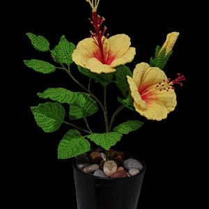 May include: A crocheted yellow hibiscus plant with red centres in a black pot with small grey stones.