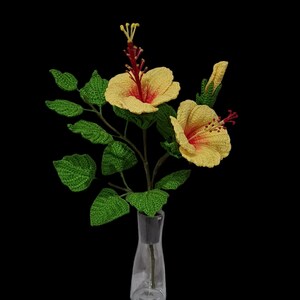 May include: A bouquet of crocheted yellow hibiscus flowers with red centres in a clear glass vase. The flowers are arranged with green crocheted leaves.