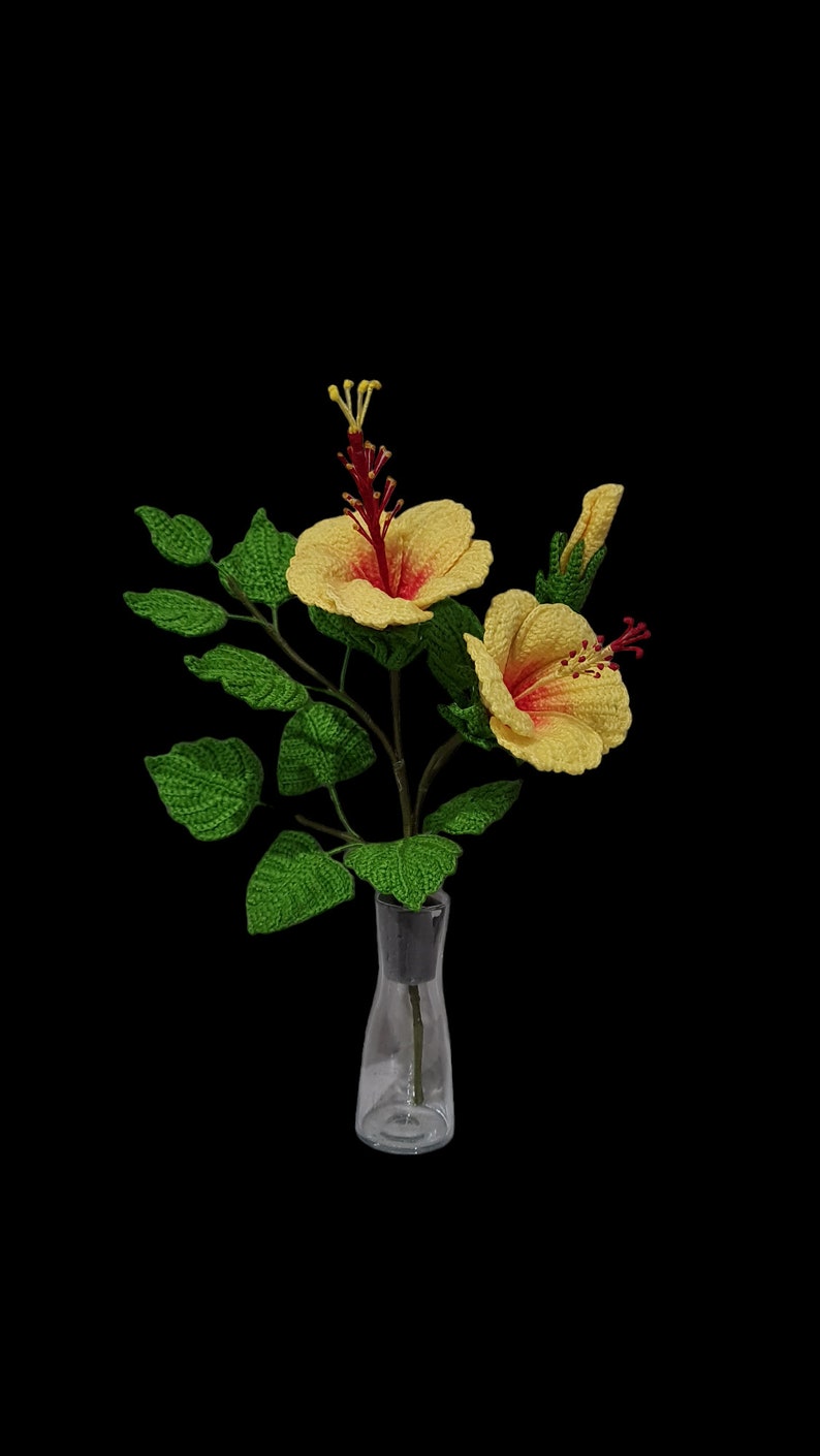 May include: A bouquet of crocheted yellow hibiscus flowers with red centres in a clear glass vase. The flowers are arranged with green crocheted leaves.