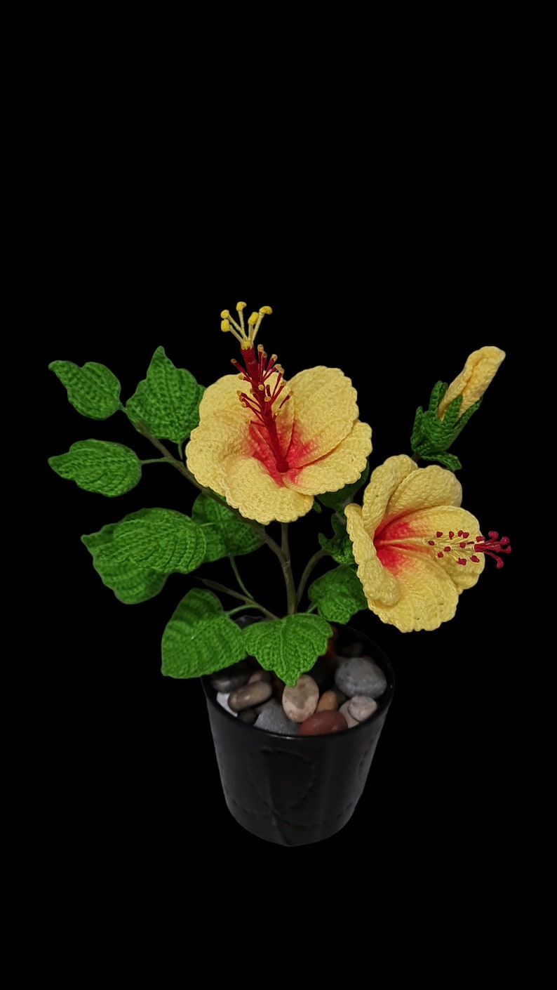 May include: A crocheted yellow hibiscus flower with a red centre in a black pot with small stones. The plant has green leaves.
