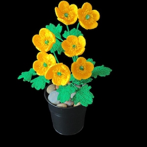 May include: A handcrafted floral arrangement featuring yellow crocheted flowers with green centers and leaves. The flowers are arranged in a black pot filled with small stones, set against a black background.