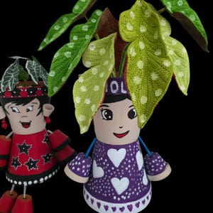 May include: Two decorative flower pots shaped like dolls. One doll wears a red dress with black stars, the other a purple dress with white hearts. Both have painted faces and hold plants. The doll on the right has the word "OL" on its head.