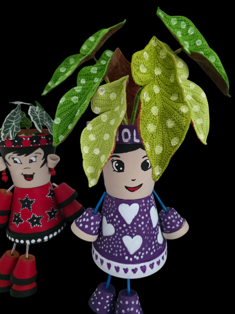 May include: Two decorative flower pots shaped like dolls. One doll wears a red dress with black stars, the other a purple dress with white hearts. Both have painted faces and hold plants. The doll on the right has the word "OL" on its head.