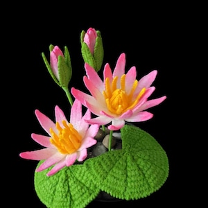 Puede incluir: Arreglo de crochet hecho a mano con nenúfares rosas y amarillos, hojas verdes y capullos. Las flores tienen pétalos rosas y centros amarillos. El fondo es negro.