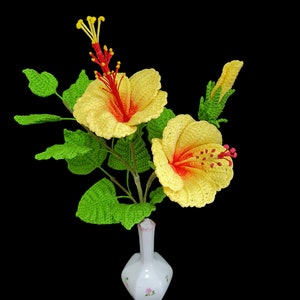 May include: A crocheted bouquet of yellow hibiscus flowers with red centres in a white vase with pink floral accents.