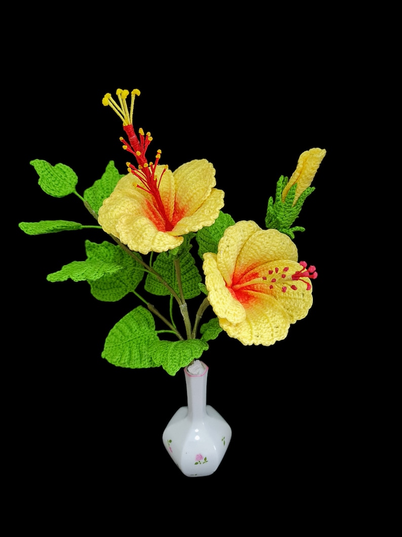 May include: A crocheted bouquet of yellow hibiscus flowers with red centres in a white vase with pink floral accents.