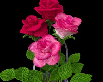 May include: A handcrafted crochet floral arrangement featuring four roses in varying shades of pink and red, with green leaves and stems. The flowers are arranged in a lavender basket against a black background.