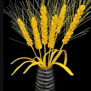 Op de afbeelding: Een boeket gele tarwehalmen gerangschikt in een donkergrijze gestreepte vaas. De tarwehalmen zijn in een waaier vorm gerangschikt met de toppen van de halmen naar boven gericht.
