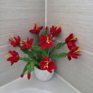 May include: A handmade crochet flower arrangement featuring vibrant red flowers with yellow stamens and green leaves, displayed in a small white ceramic pot. The decorative floral piece is placed in a corner against a gray tiled wall.