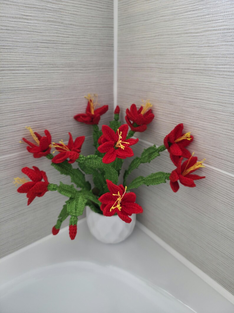 May include: A handmade crochet flower arrangement featuring vibrant red flowers with yellow stamens and green leaves, displayed in a small white ceramic pot. The decorative floral piece is placed in a corner against a gray tiled wall.