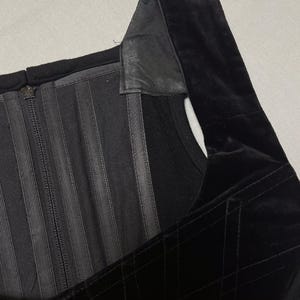 May include: Close-up of a black corset top. The top has a square neckline, a velvet bodice with a quilted pattern, and a zip closure. The straps are made of a different material, possibly leather.