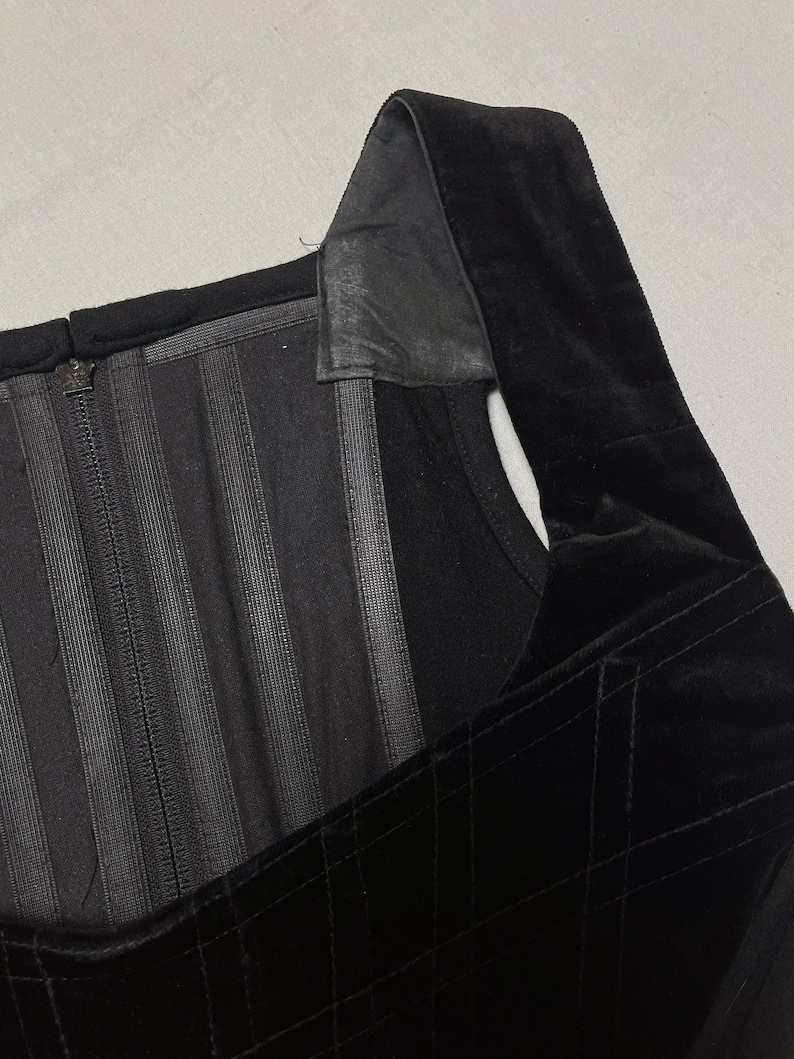May include: Close-up of a black corset top. The top has a square neckline, a velvet bodice with a quilted pattern, and a zip closure. The straps are made of a different material, possibly leather.