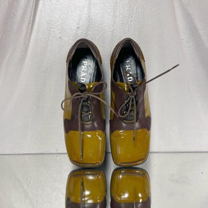 May include: A pair of two-tone leather shoes. The shoes are brown and mustard yellow with laces. The word "PRADA" is visible on the insole of each shoe. The shoes are reflected on a shiny surface.