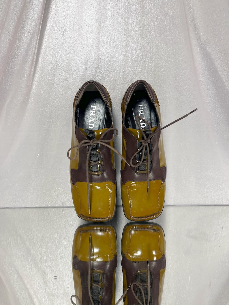 May include: A pair of two-tone leather shoes. The shoes are brown and mustard yellow with laces. The word "PRADA" is visible on the insole of each shoe. The shoes are reflected on a shiny surface.