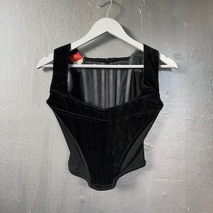 May include: A black velvet corset top with a square neckline and structured bodice. The corset is hanging on a white hanger against a grey background. The top features vertical paneling and a zip closure at the back.