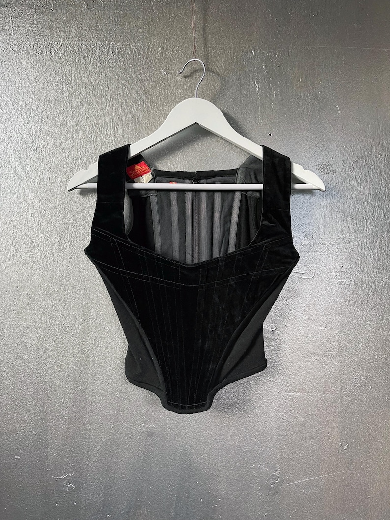 May include: A black velvet corset top with a square neckline and structured bodice. The corset is hanging on a white hanger against a grey background. The top features vertical paneling and a zip closure at the back.