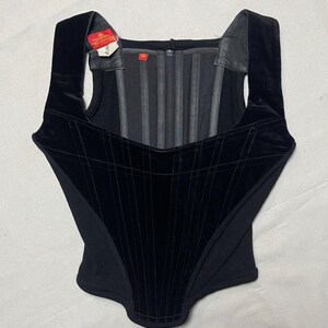 May include: A black corset with a square neckline and wide shoulder straps. The corset features a velvet panel with vertical and horizontal stitching, creating a grid pattern. A tag is attached to the strap. The corset has a zip closure.