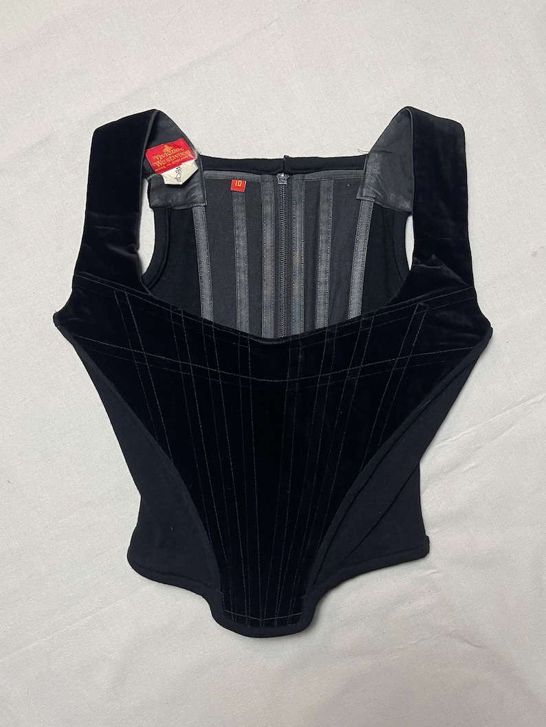 May include: A black corset with a square neckline and wide shoulder straps. The corset features a velvet panel with vertical and horizontal stitching, creating a grid pattern. A tag is attached to the strap. The corset has a zip closure.