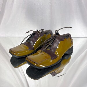 May include: A pair of two-tone, olive green and brown leather shoes with square toes and lace-up closures. The shoes are reflected in a mirrored surface, showcasing their glossy finish and design.