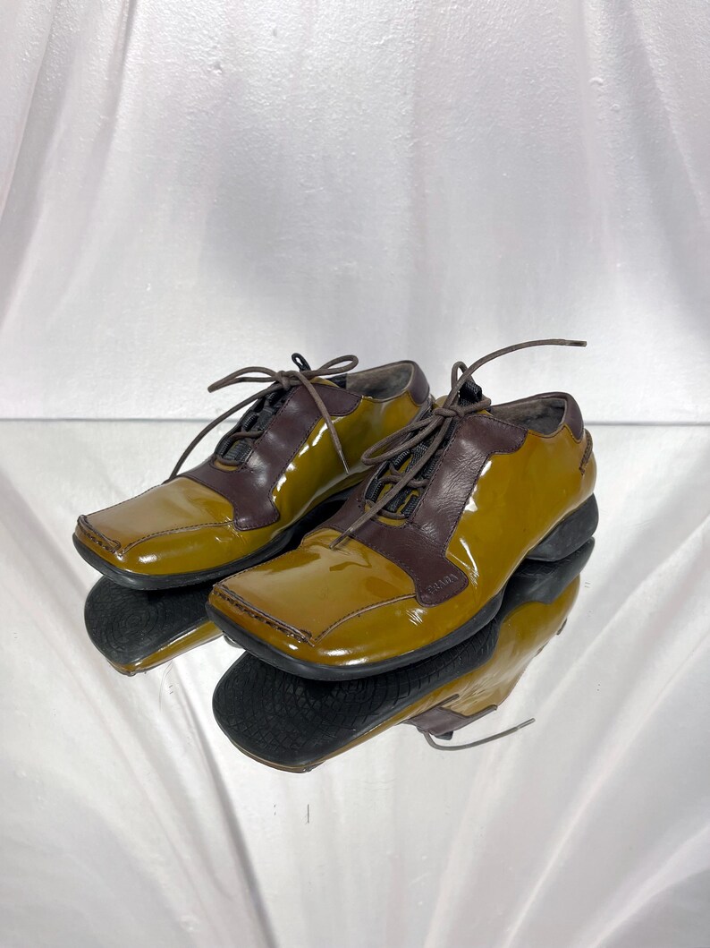 May include: A pair of two-tone, olive green and brown leather shoes with square toes and lace-up closures. The shoes are reflected in a mirrored surface, showcasing their glossy finish and design.
