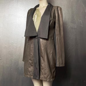 Puede incluir: Un abrigo largo de cuero marrón con cuello y solapas negras. El abrigo tiene cierre de botón y un corte entallado. Las mangas son largas y el abrigo llega hasta la mitad del muslo. El abrigo se exhibe en un maniquí.