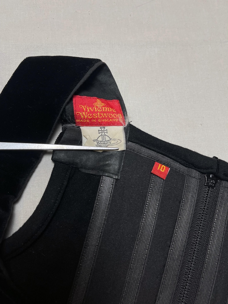May include: Close-up of a black garment with a velvet strap. A red tag with "Vivienne Westwood" and "MADE IN ENGLAND" is visible. The garment has vertical striped detailing and a size 10 tag.