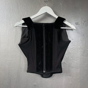 May include: Black corset top with a square neckline and velvet detailing. The top has a central zip and is suspended on a white hanger against a grey backdrop. The straps are made of velvet.