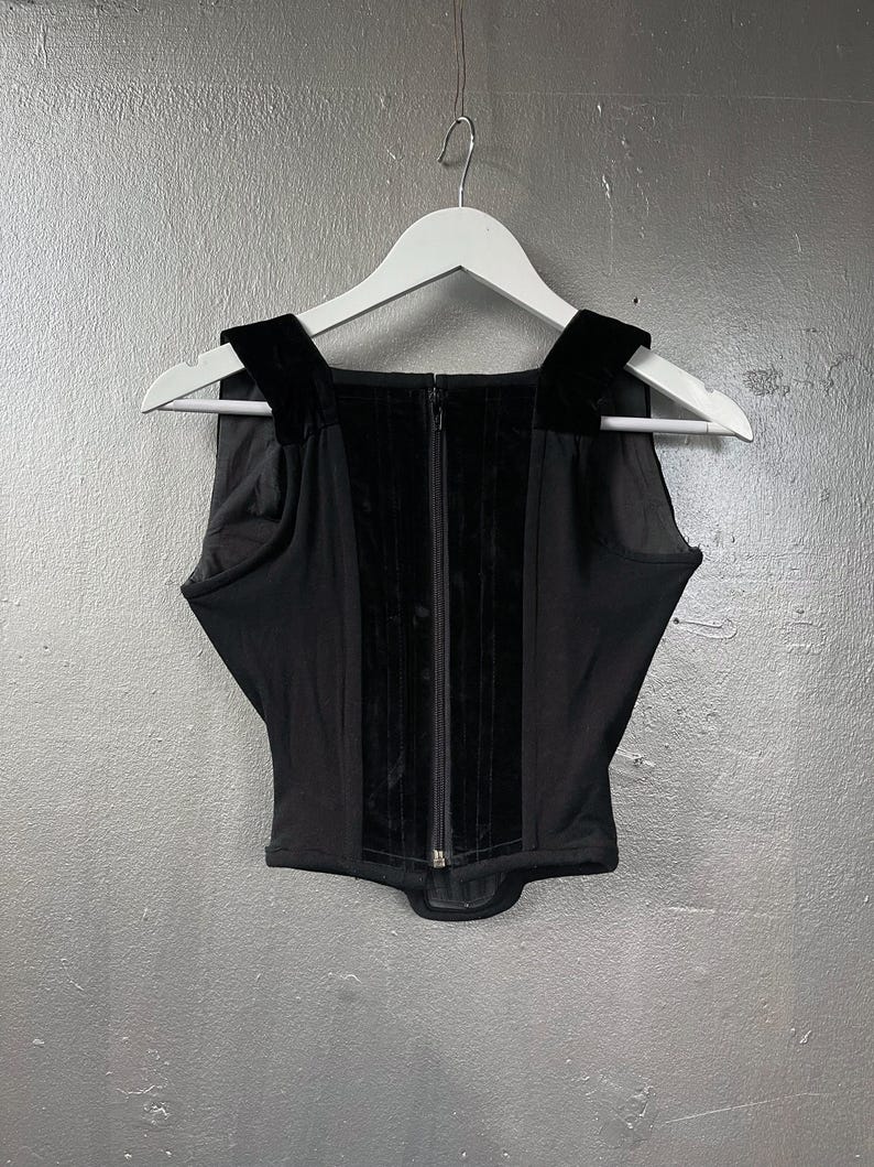 May include: Black corset top with a square neckline and velvet detailing. The top has a central zip and is suspended on a white hanger against a grey backdrop. The straps are made of velvet.