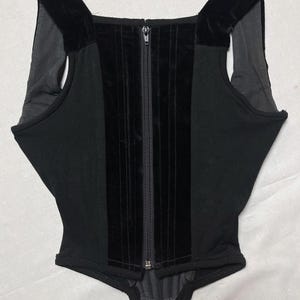 May include: Black corset top with a front zip and velvet detailing. The top has a square neckline and velvet shoulder straps. The central panel is velvet, and the side panels are a matte black fabric. A stylish garment.