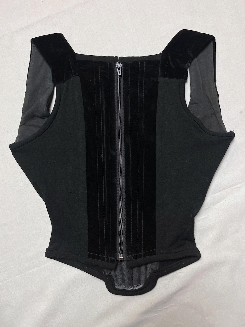 May include: Black corset top with a front zip and velvet detailing. The top has a square neckline and velvet shoulder straps. The central panel is velvet, and the side panels are a matte black fabric. A stylish garment.