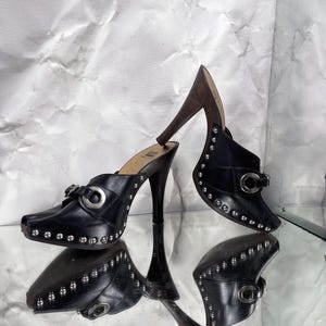 May include: Black leather high-heeled clogs with silver-tone stud accents and buckles. The heels are dark brown and the shoes are reflected on a mirrored surface. The clogs have a pointed toe.