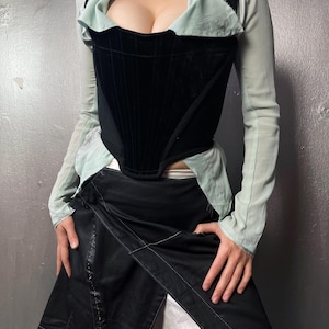 May include: A black velvet corset with a structured design, worn with a light green collared top and long sleeves. A black wrap skirt with white stitching and white trousers complete the outfit.
