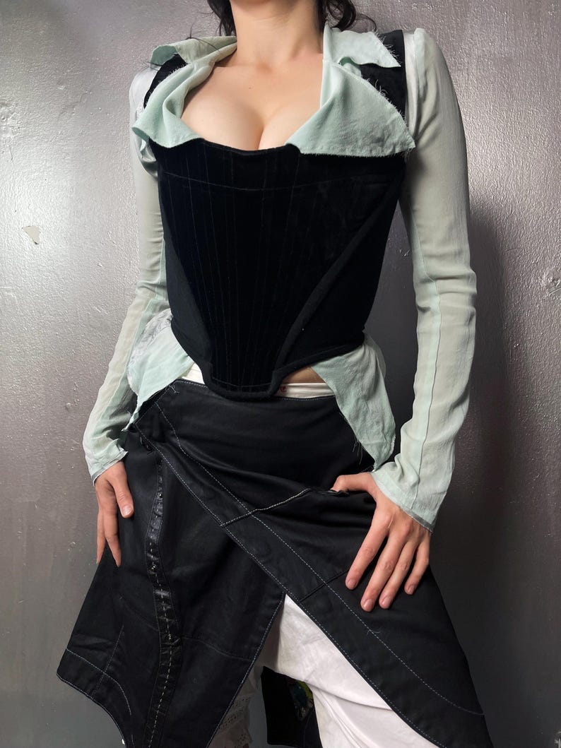 May include: A black velvet corset with a structured design, worn with a light green collared top and long sleeves. A black wrap skirt with white stitching and white trousers complete the outfit.
