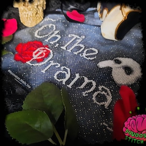 May include: A denim-textured fabric with the phrase "Oh, The Drama" embroidered in white. A red rose, a skull, and a Phantom of the Opera mask are arranged around the text. The image has a dark, theatrical aesthetic.