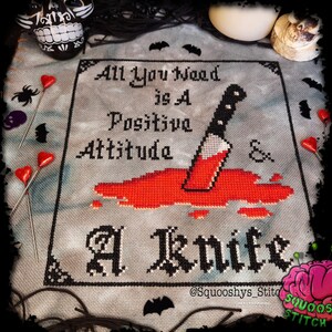 May include: Cross stitch pattern with a black and white design of a knife with red blood dripping from it. The text reads "All you need is a positive attitude & a knife".