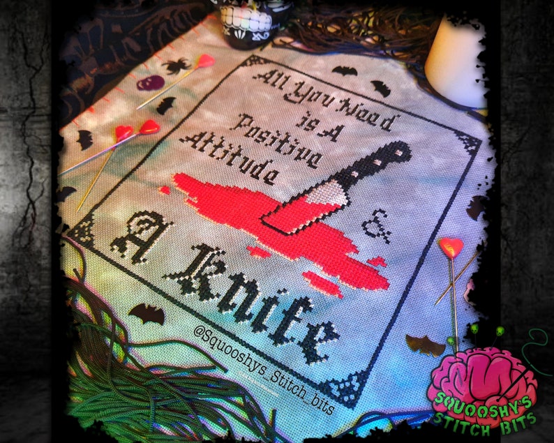 May include: Cross stitch pattern with a bloody knife and the text "All you need is a positive attitude & a knife" with the artist's social media handle "@squoooshys_stitch_bits" at the bottom.