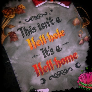 Könnte beinhalten: Kreuzstich-Stickerei mit grauem Hintergrund und schwarzem und orangefarbenem Text, der "This isn't a Hell-hole It's a Hell-home" lautet.