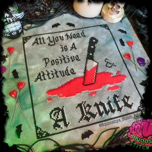 May include: Cross stitch pattern with a black and white design that says "All You Need is A Positive Attitude & A Knife". The design is on a light blue background with red blood splatters around a black knife.