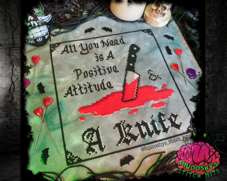 May include: Cross stitch pattern with a black and white design that says "All You Need is A Positive Attitude & A Knife". The design is on a light blue background with red blood splatters around a black knife.