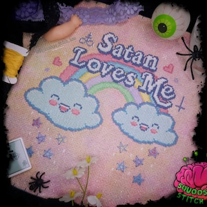 May include: A cross-stitch design with the text "Satan Loves Me" above a rainbow and two smiling cloud characters. The design is on a pink background with stars and hearts. The image includes a toy eyeball, a spider, and a spool of yellow thread.